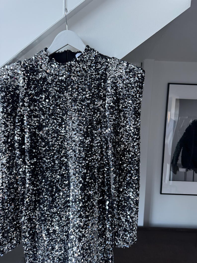 May include: A black dress covered in silver sequins hangs on a white hanger. The dress has long sleeves and a high neckline. The sequins create a shimmering effect, reflecting light. The dress is a formal garment.