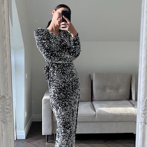 May include: A woman wearing a long, black sequined dress with a silver shimmer. The dress has long sleeves and a fitted silhouette. The woman is standing in front of a mirror, and a couch is visible in the background.