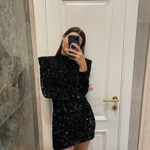 May include: A black sequined dress with long sleeves and a high neckline. The dress is a mini-skirt style, reflecting light. The garment is suitable for special occasions and evening wear.