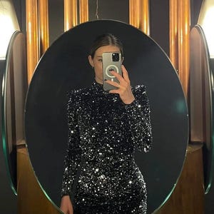 May include: A black sequin dress with long sleeves and a high neckline is reflected in an oval mirror. The dress sparkles with silver sequins. The mirror is framed by a wooden border, and the background features copper-colored columns.