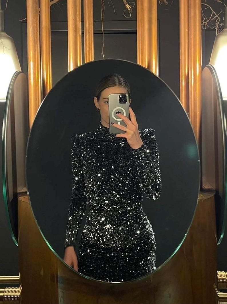 May include: A black sequin dress with long sleeves and a high neckline is reflected in an oval mirror. The dress sparkles with silver sequins. The mirror is framed by a wooden border, and the background features copper-colored columns.