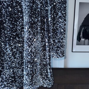 May include: A black dress covered in silver sequins. The long-sleeved dress has a high neckline and a slightly flared skirt. The sequins catch the light, creating a shimmering effect. The dress is hanging against a white wall.