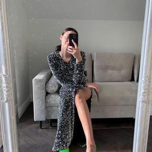 May include: A woman wearing a black sequined dress and bright green high heels. She is sitting on a gray couch in front of a mirror.