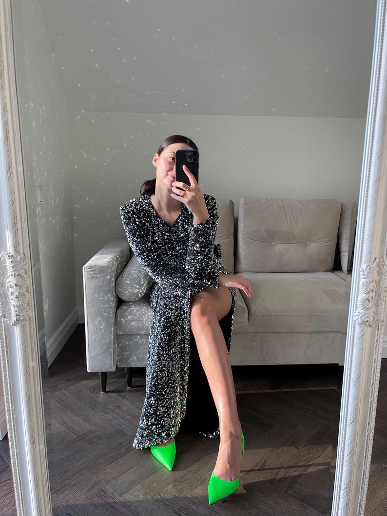 May include: A woman wearing a black sequined dress and bright green high heels. She is sitting on a gray couch in front of a mirror.