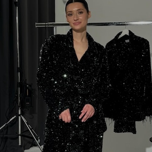 May include: A woman wearing a black sequined blazer and matching pants. The blazer has a V-neck and is paired with a similar sequined jacket hanging on a rack. The outfit sparkles under the studio lights, creating a glamorous look.