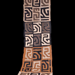 Peut inclure: Un long chemin de table textile rectangulaire présentant une grille répétitive de panneaux carrés. Chaque panneau affiche un motif géométrique abstrait unique dans des tons de noir, brun foncé, beige et crème, créé par des coutures complexes.