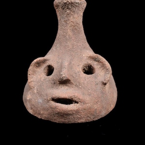 May include: A brown, textured clay vessel shaped like a human head with a narrow neck opening at the top. The face features two circular eye cutouts, a small protruding nose, and an open, oval-shaped mouth, all crafted from rough, earthy material.