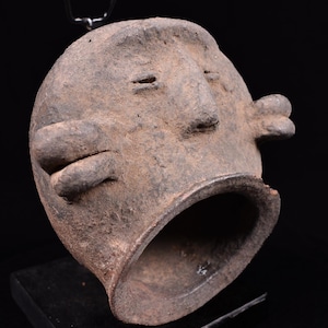 May include: A dark brown, textured ceramic vessel shaped like a stylized human head. The object features a wide, circular opening at the base, a prominent nose, two horizontal slit eyes, and two protruding, rounded ear-like appendages on the sides.