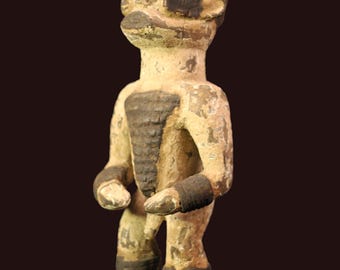May include: A weathered, tan-colored wooden figurine of a standing human figure. The figure has a dark brown headdress, chest plate, and bands around the wrists and ankles. The figure's face has simple features, and the overall style suggests an antique or tribal art piece.