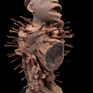 Puede incluir: Una figura antropomórfica de madera tallada con sombrero, que presenta una cavidad en el torso y numerosos clavos de hierro oxidados incrustados por todo el cuerpo, sobre una base circular de madera con fondo negro.