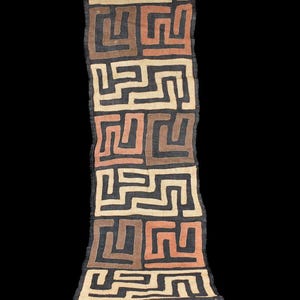 Puede incluir: Textil rectangular y largo con un patrón geométrico repetitivo en tonos marrones, beige y negro. El diseño presenta formas entrelazadas, creando una apariencia laberíntica. La tela tiene un borde ligeramente deshilachado, lo que sugiere calidad artesanal.