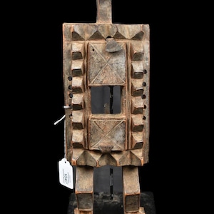 Puede incluir: Escultura de madera tallada de una figura de pie con cuerpo rectangular y cabeza triangular. La figura presenta patrones geométricos y un aspecto envejecido, montada sobre una base negra. Probablemente sea una obra de arte tribal africana.