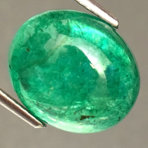 Pode incluir: Uma esmeralda oval de cor verde vibrante com uma superf&iacute;cie lisa e polida. A pedra tem um aspecto ligeiramente nublado com inclus&otilde;es vis&iacute;veis.