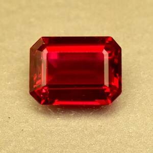 May include: A faceted, rectangular-cut ruby gemstone. The gem is a deep, saturated red color with sharp, clean edges. The gemstone is set against a neutral background, highlighting its brilliance and clarity. This gemstone is suitable for jewelry making.
