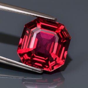 May include: A radiant-cut red gemstone held by tweezers. The gem's deep red colour is contrasted by a lighter, reflective interior. The facets create a geometric pattern, reflecting light. The background is a dark grey.