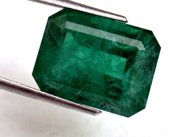 May include: A vibrant green emerald gemstone with a rectangular cut, held by tweezers against a white background.