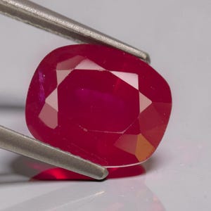 Puede incluir: Una gema de rubí tallada en cojín, sujeta con pinzas plateadas. El rubí es de un color rojo intenso y saturado con múltiples facetas que reflejan la luz. La gema está sobre un fondo blanco.