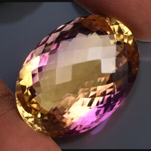 May include: An oval-cut gemstone displaying a gradient of colours. The gem transitions from a golden-yellow to a pinkish-purple hue. The facets create multiple reflective surfaces, enhancing its brilliance and sparkle.