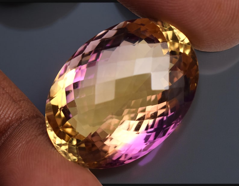 May include: An oval-cut gemstone displaying a gradient of colours. The gem transitions from a golden-yellow to a pinkish-purple hue. The facets create multiple reflective surfaces, enhancing its brilliance and sparkle.