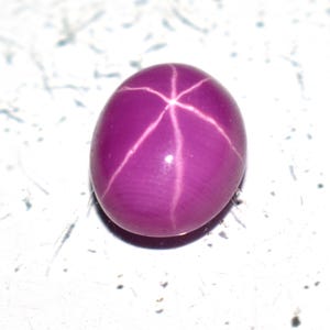 Puede incluir: Una gema pulida, de forma ovalada, de color rojo púrpura, con un patrón de estrella de seis rayos en su superficie. El efecto de estrella se crea por la luz que se refleja en las inclusiones dentro de la piedra. La gema está sobre un fondo blanco.
