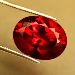 Könnte beinhalten: Ein facettierter, ovalförmiger, tiefroter Edelstein wird von einer silbernen Pinzette gehalten. Die Oberfläche des Edelsteins reflektiert das Licht und zeigt seine vielen Facetten und seine intensive Farbe. Der Hintergrund ist ein warmes Beige.
