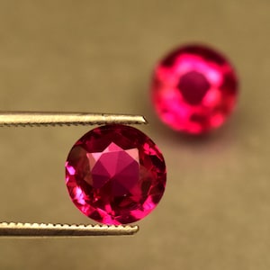May include: Close-up of two faceted, round, deep pink gemstones. One is held by tweezers, showcasing its intricate facets. The other gem is slightly out of focus in the background. The gems are set against a neutral background.
