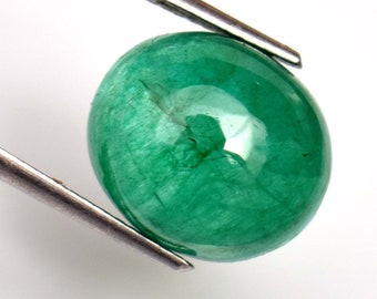 Pode incluir: Um cabochão de esmeralda verde vibrante, com uma superfície lisa e arredondada. A pedra é mantida no lugar por uma pinça.