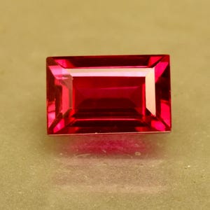 Könnte beinhalten: Ein rechteckiger, facettierter, rubinroter Edelstein. Der Edelstein hat eine glänzende, polierte Oberfläche mit scharfen Kanten und eine symmetrische Form. Der Edelstein ist vor einem neutralen Hintergrund platziert, der seine lebendige Farbe und Klarheit hervorhebt.