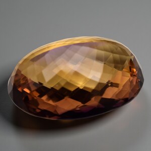 May include: A faceted, oval gemstone displaying a colour gradient. The gem transitions from a deep reddish-brown at the base to a golden-yellow hue at the top, with hints of purple. The cut of the stone creates multiple reflective surfaces.