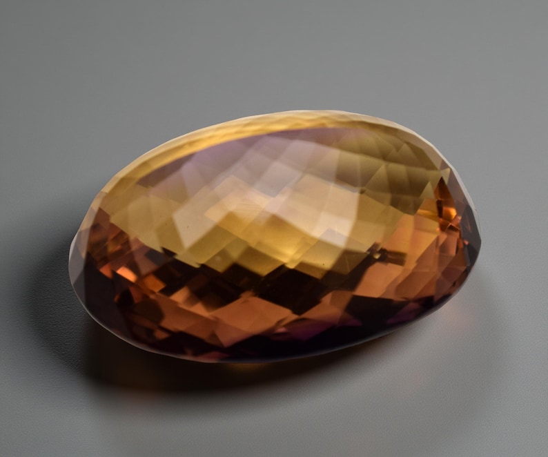 May include: A faceted, oval gemstone displaying a colour gradient. The gem transitions from a deep reddish-brown at the base to a golden-yellow hue at the top, with hints of purple. The cut of the stone creates multiple reflective surfaces.