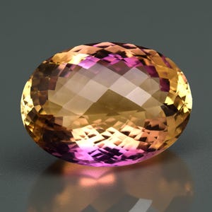 May include: An oval-shaped gemstone with a faceted surface. The gem exhibits a colour gradient, shifting from golden yellow to a vibrant pinkish-purple. The stone is set against a neutral grey background, accentuating its brilliance.