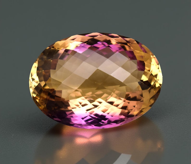 May include: An oval-shaped gemstone with a faceted surface. The gem exhibits a colour gradient, shifting from golden yellow to a vibrant pinkish-purple. The stone is set against a neutral grey background, accentuating its brilliance.