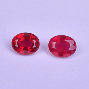 Puede incluir: Dos piedras preciosas rojas de forma ovalada con corte facetado. Las piedras preciosas están sobre un fondo blanco.