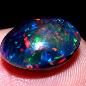 May include: A dark grey oval-shaped opal gemstone with a black background. The opal has a rainbow of colours including red, green, blue, and purple.