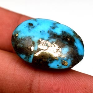 May include: Oval shaped turquoise gemstone with gold flecks and black matrix. The stone is a vibrant blue colour.