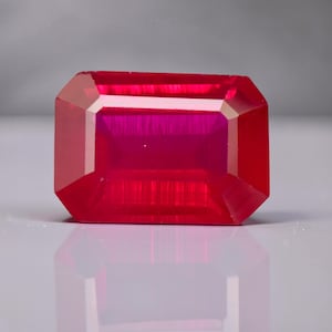 May include: A vibrant, rectangular-cut ruby gemstone. The gem is a deep, saturated red with a polished surface, reflecting light and showcasing its facets. The ruby's shape is an emerald cut, with a clear, internal structure.
