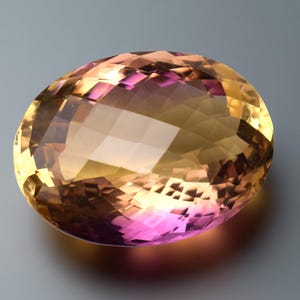 May include: An oval-cut gemstone displaying a colour gradient. The gem transitions from a golden yellow to a pinkish-purple hue. The facets create multiple reflective surfaces, enhancing its brilliance and sparkle.