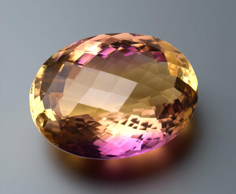 May include: An oval-cut gemstone displaying a colour gradient. The gem transitions from a golden yellow to a pinkish-purple hue. The facets create multiple reflective surfaces, enhancing its brilliance and sparkle.