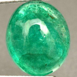 Pode incluir: Uma gema de esmeralda verde vibrante, cortada em forma oval, com uma superf&iacute;cie lisa e polida. A pedra &eacute; segurada por uma pin&ccedil;a.