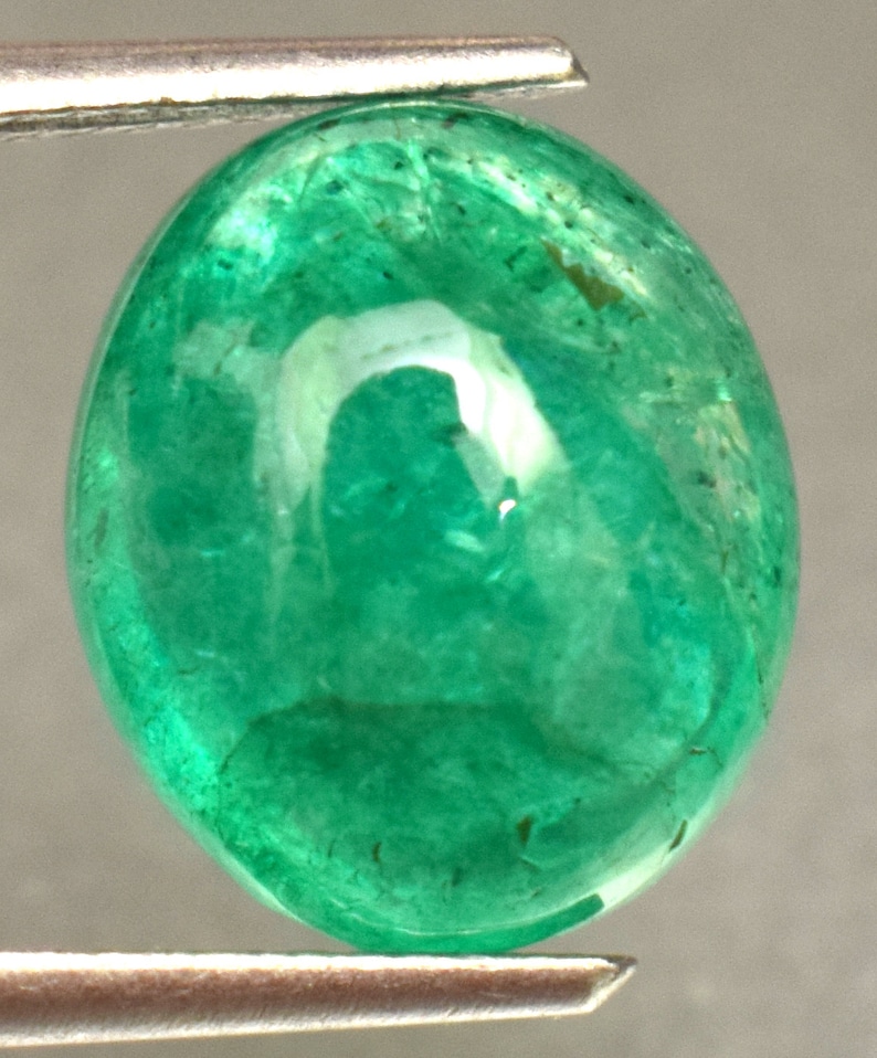 Pode incluir: Uma gema de esmeralda verde vibrante, cortada em forma oval, com uma superf&iacute;cie lisa e polida. A pedra &eacute; segurada por uma pin&ccedil;a.