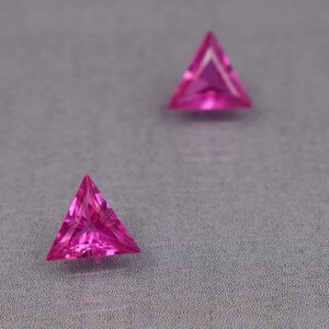 Puede incluir: Dos piedras preciosas triangulares de color rosa sobre una superficie gris texturizada.