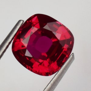 Könnte beinhalten: Ein leuchtend roter Edelstein mit einem Kissen-Schliff. Der Edelstein wird in einer Pinzette gehalten.