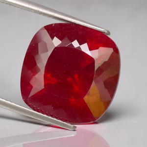 Puede incluir: Una gema de rubí facetada con corte cojín, sujeta con pinzas plateadas. La gema es de un color rojo intenso y saturado, con múltiples facetas que reflejan la luz. La forma es un cuadrado redondeado.