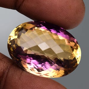 May include: An oval-cut gemstone with a blend of yellow and purple hues. The faceted surface reflects light, creating a sparkling effect. The gem is held between fingers, showcasing its size and colour variations.
