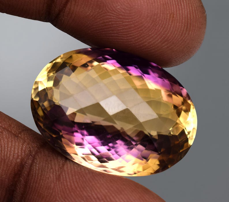 May include: An oval-cut gemstone with a blend of yellow and purple hues. The faceted surface reflects light, creating a sparkling effect. The gem is held between fingers, showcasing its size and colour variations.