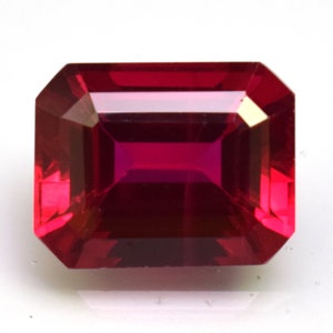 May include: A deep red gemstone with a rectangular emerald cut. The gemstone is faceted and has a high polish.