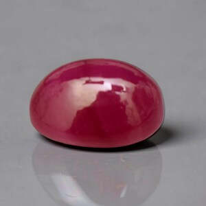 May include: A smooth, oval-shaped cabochon gemstone with a deep, saturated magenta-red color. The surface is highly polished, creating a reflective, glossy finish that captures light, highlighting the rounded dome and uniform, vibrant red hue.