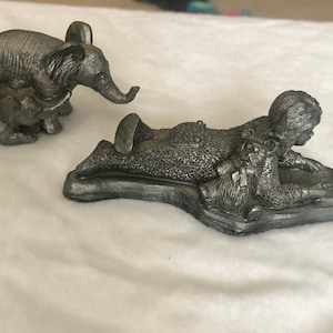 May include: Two pewter figurines, one of a mother and baby elephant, the other of a child lying down with a teddy bear. The child is holding a book and appears to be reading.