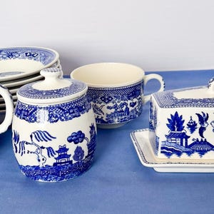 Op de afbeelding: Een blauw-wit keramisch servies met een Willow-patroon. De set bevat een melkkan, suikerpot, kopje, botervloot en borden. De stukken hebben een klassieke, vintage uitstraling.