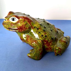 May include: A ceramic frog figurine in shades of green and reddish-brown. The frog has a textured surface with black spots and white eyes. The figurine is a decorative item.